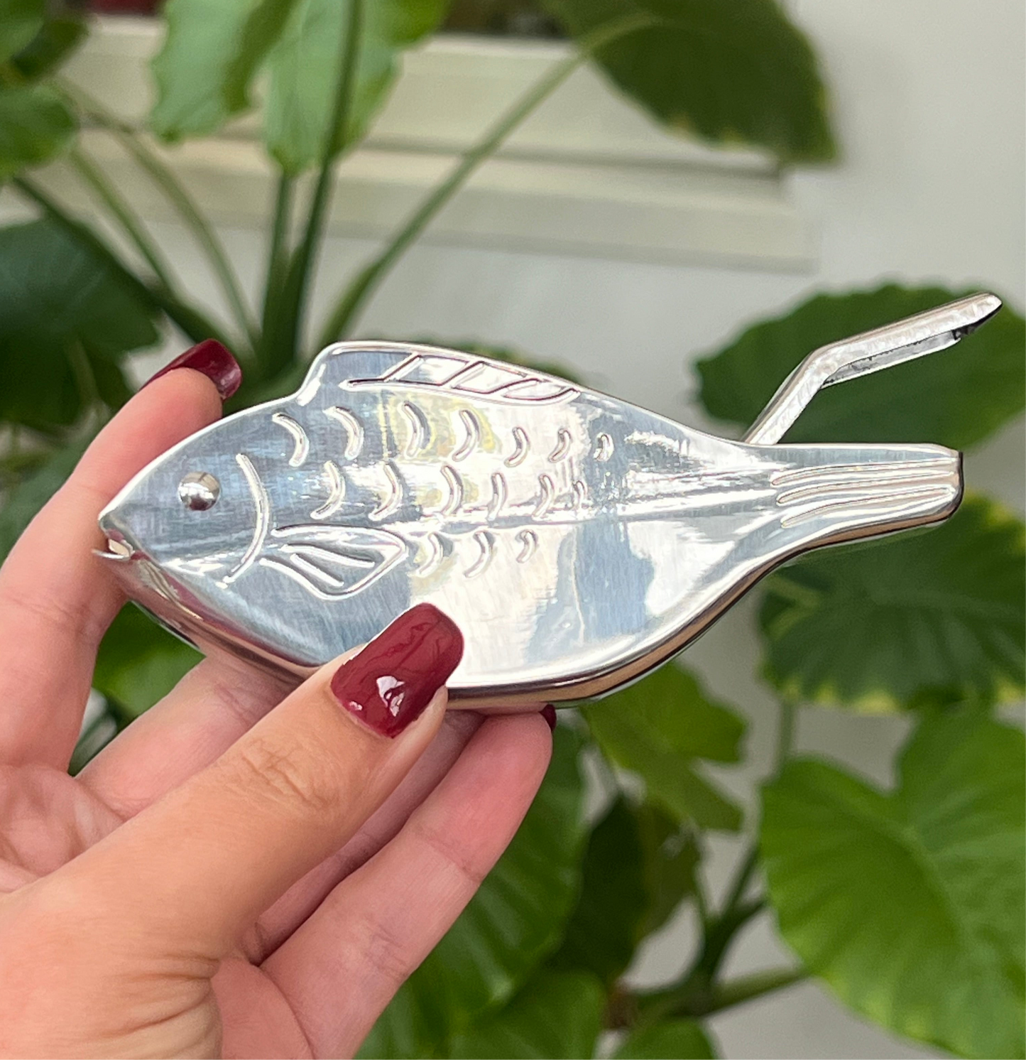 Silver fish-shaped object held by a hand with red nail polish, against a green leafy background.