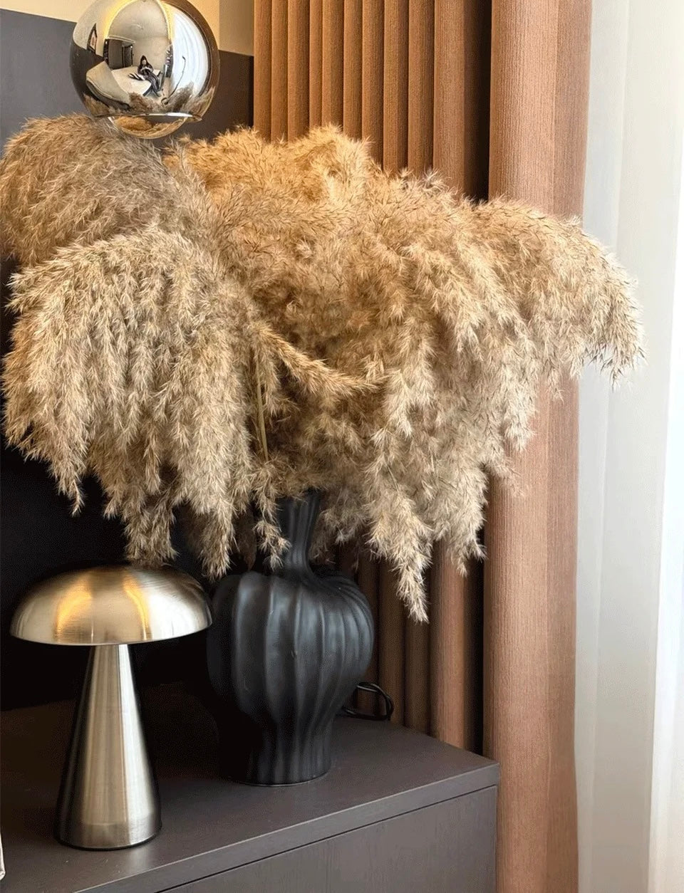 Decorative arrangement with pampas grass, a dark vase, and a metallic lamp on a shelf.