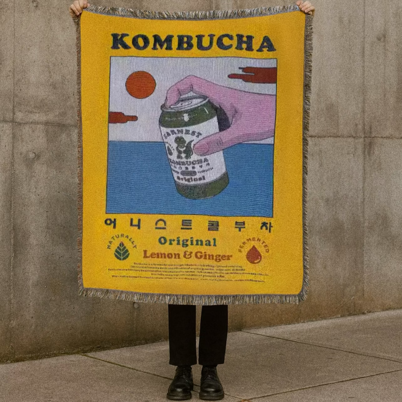 Person holding a kombucha-themed blanket against a concrete wall.