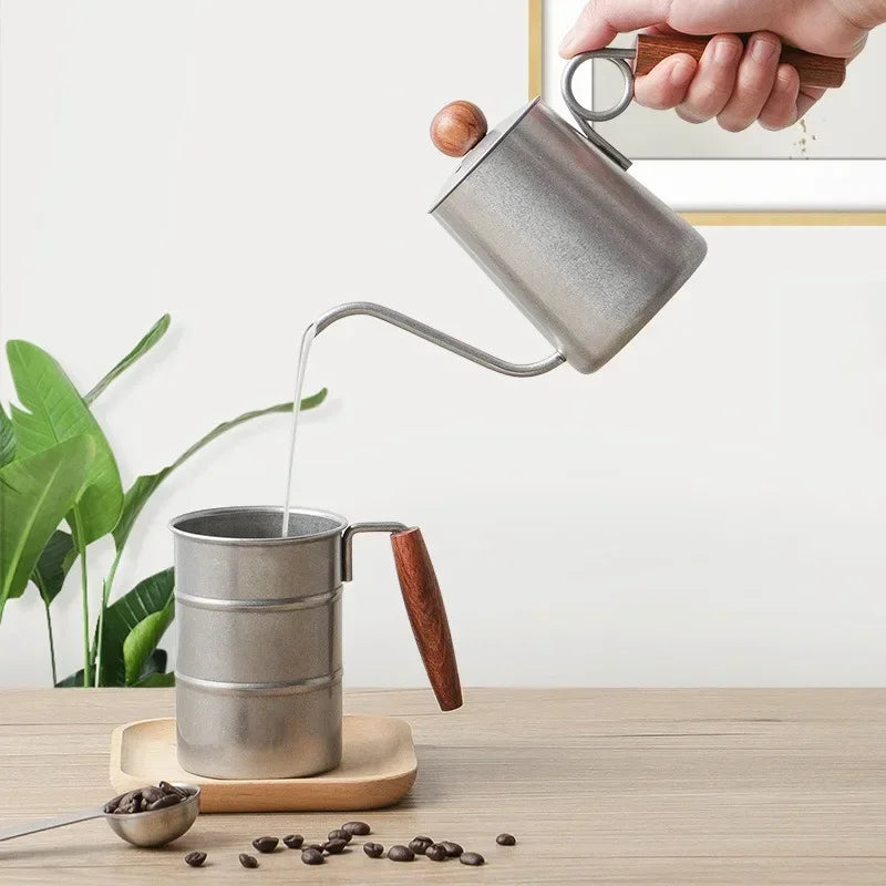 Person pouring coffee from a metal pour-over coffee maker into another container on a wooden surface with coffee beans and a plant in the background.