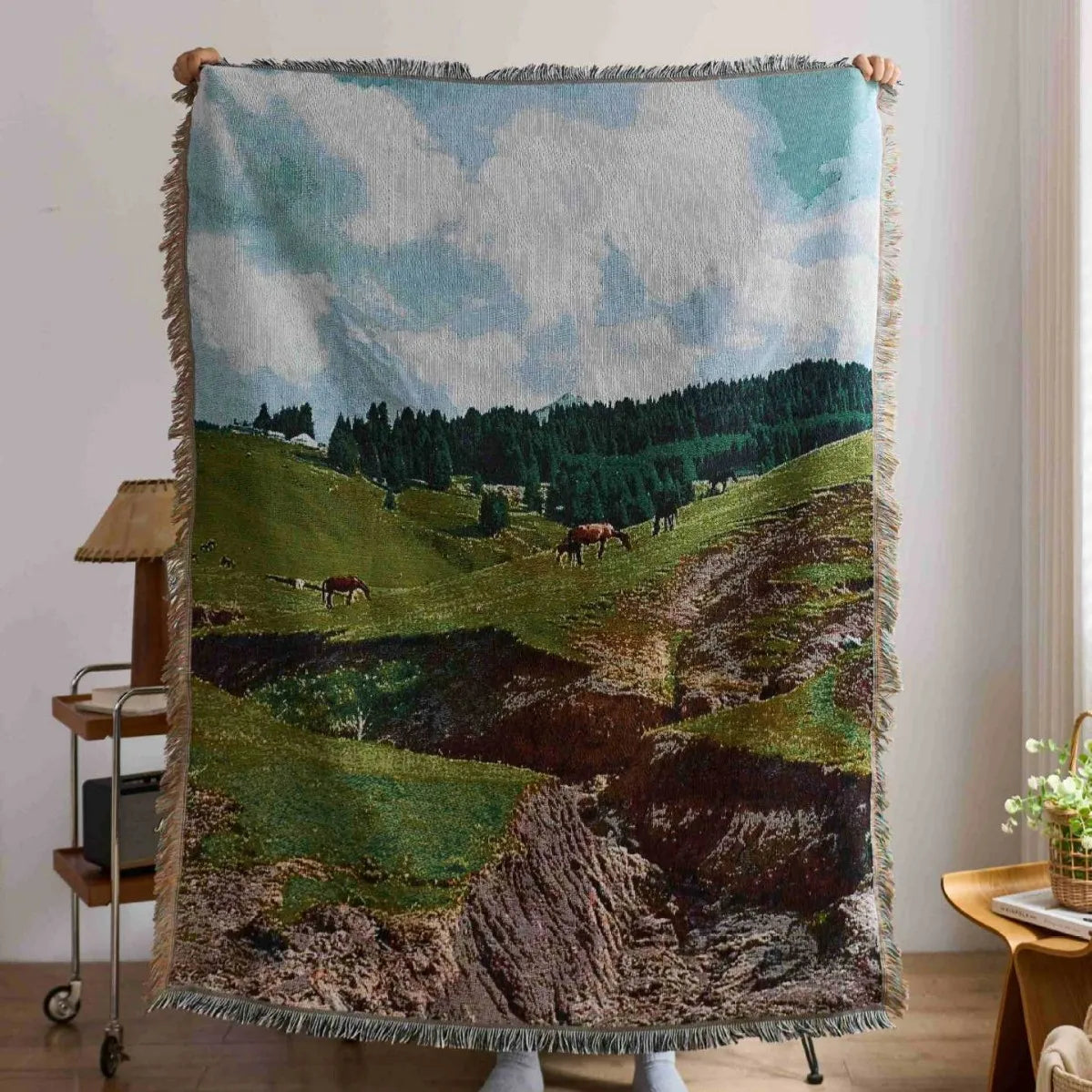Woven blanket with a landscape design of green hills and trees held up in a room.