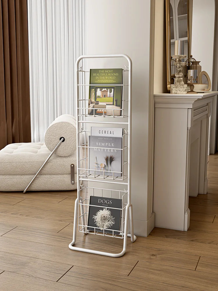 White magazine rack with books on a wooden floor in a room with a mirror and sofa.