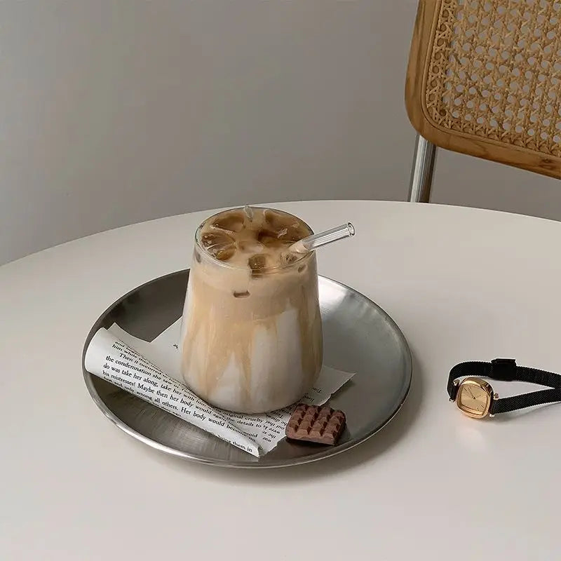 Glass of iced coffee on a metal tray with a waffle cookie and newspaper, placed on a white table with a wooden chair.