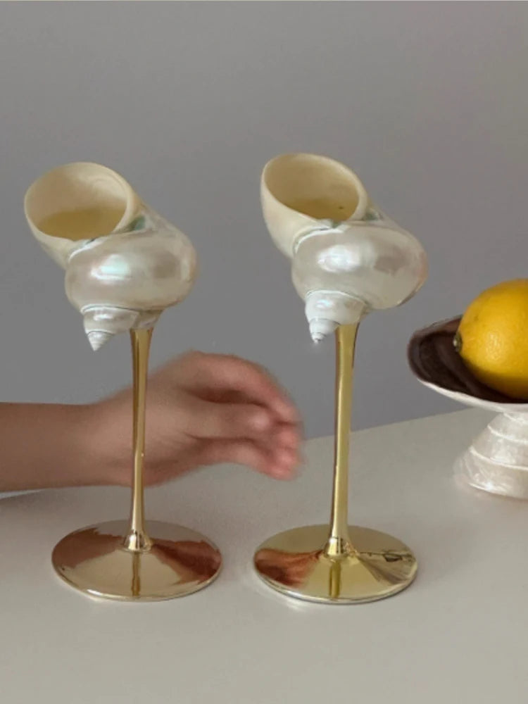 Two decorative shell-shaped candle holders on gold stands with a hand reaching towards them.