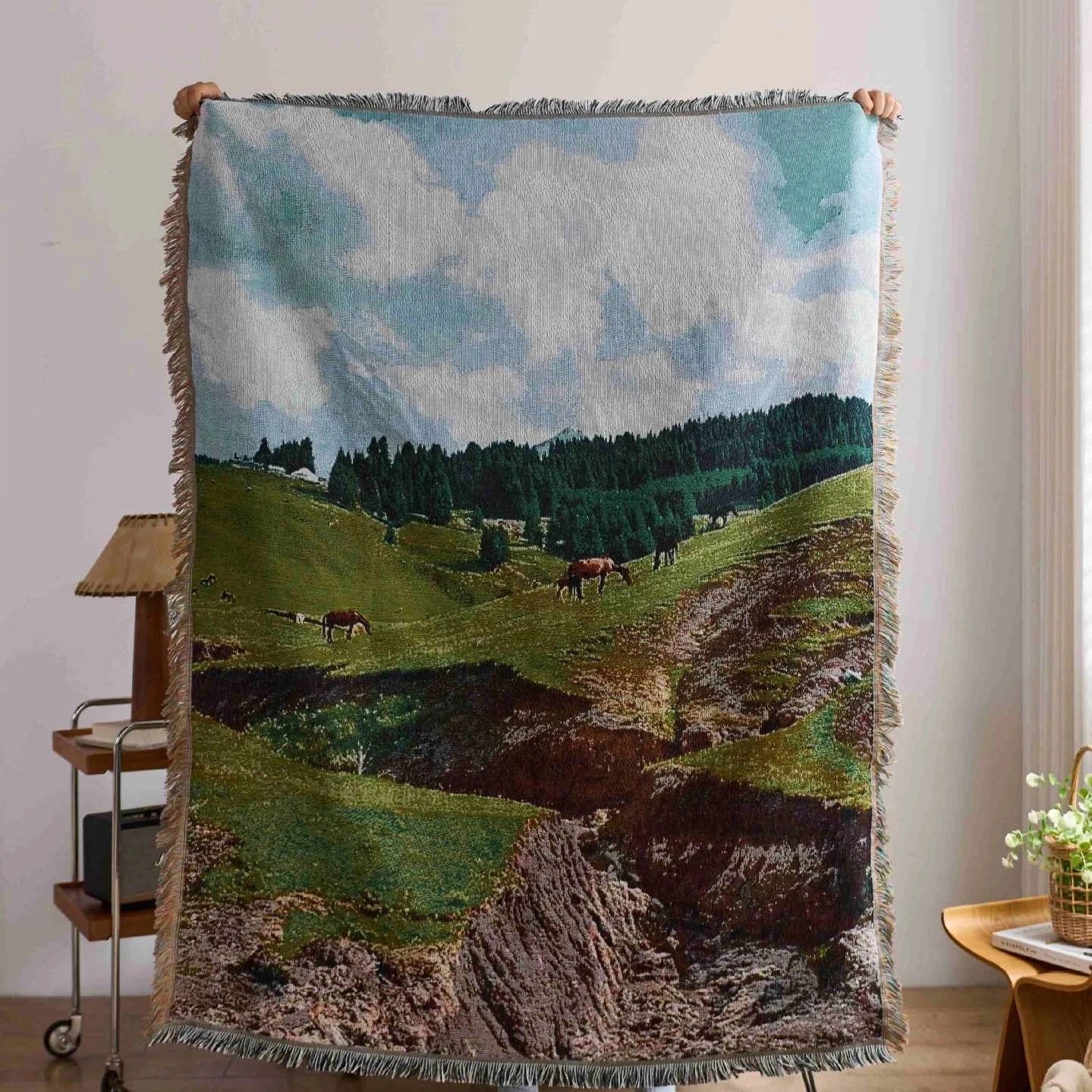 Blanket with a landscape design of cows in a field held up against a white wall.