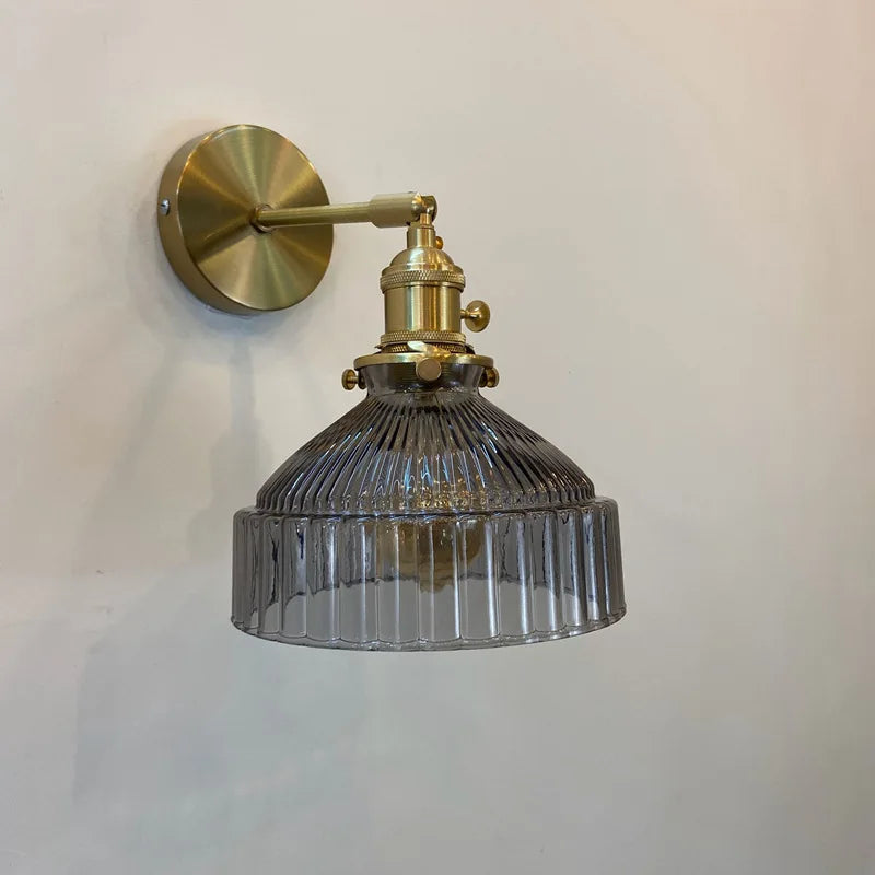 Wall-mounted light fixture with brass arm and ribbed glass shade on a plain background