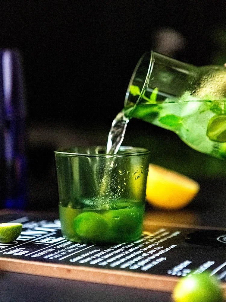 Green drink being poured into a glass with limes and a carafe in the background.