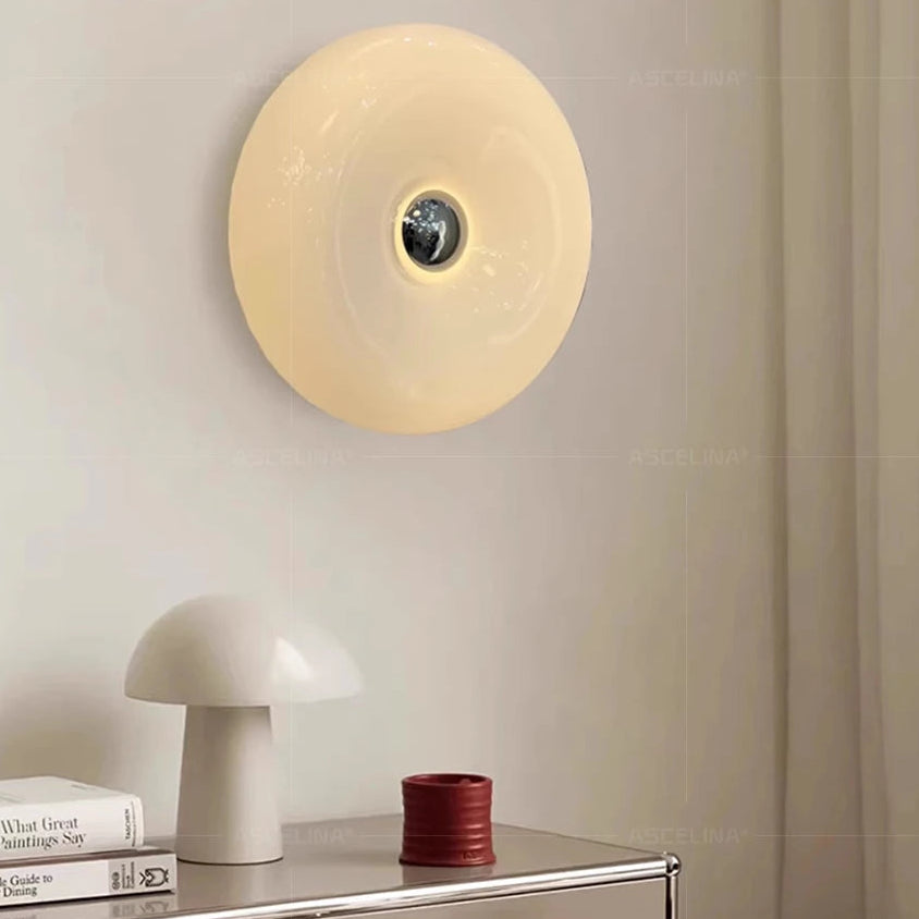 Round wall light fixture on a neutral wall with a shelf below containing books and a small container.