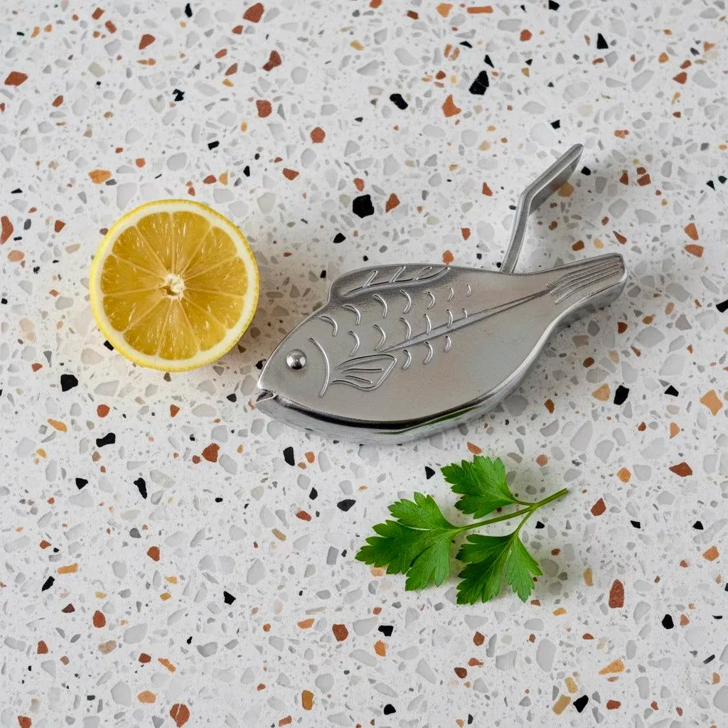 Metallic fish-shaped lemon squeezer on a speckled countertop with a lemon and parsley.