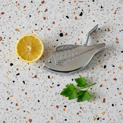 Metallic fish-shaped lemon squeezer on a speckled countertop with a lemon and parsley.