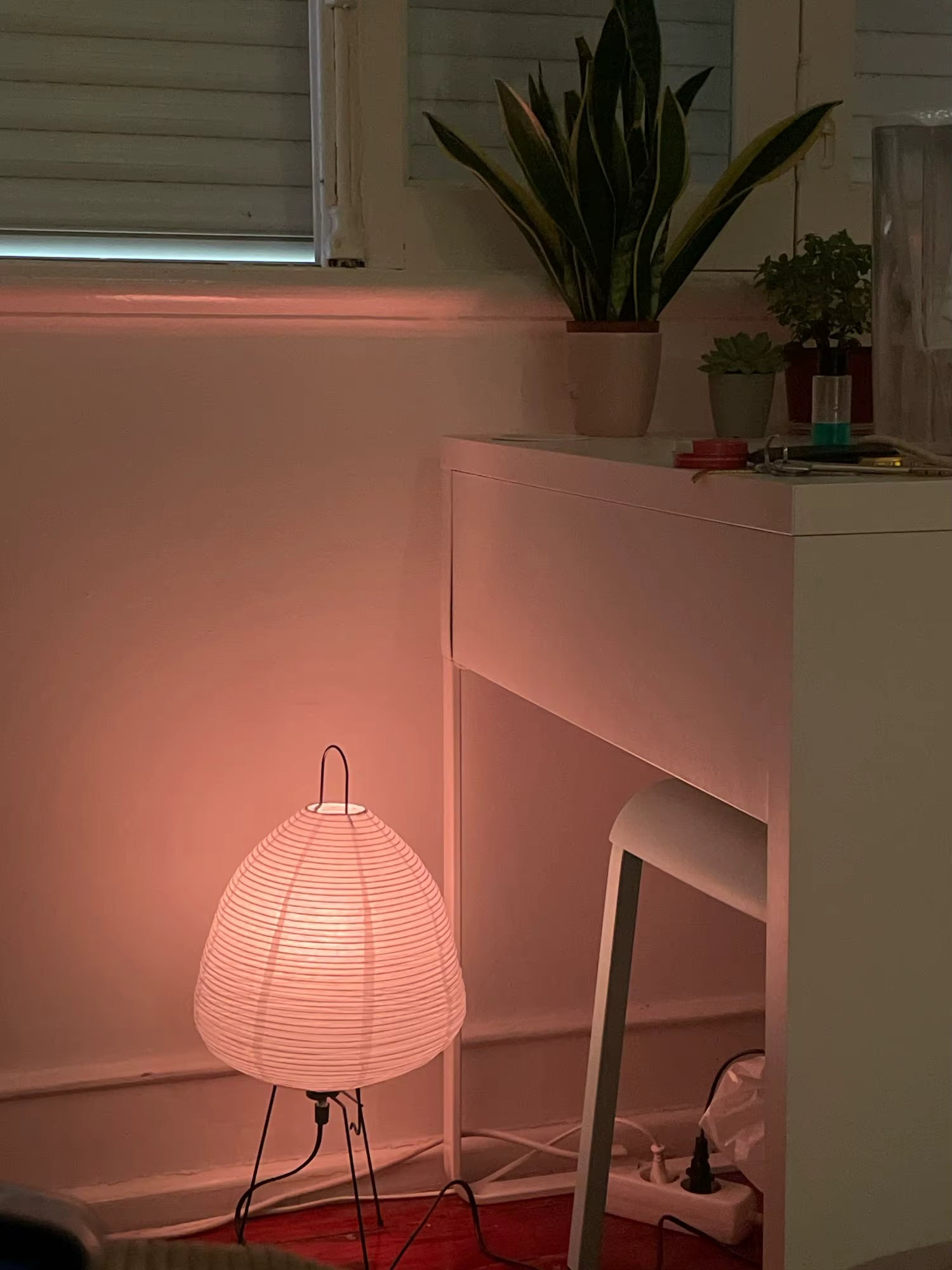 Woven lamp on a red rug next to a white cabinet with plants in the background