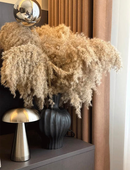 Decorative arrangement with pampas grass, a dark vase, and a metallic lamp on a shelf.