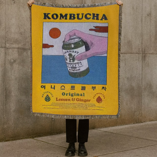 Person holding a kombucha-themed blanket against a concrete wall.