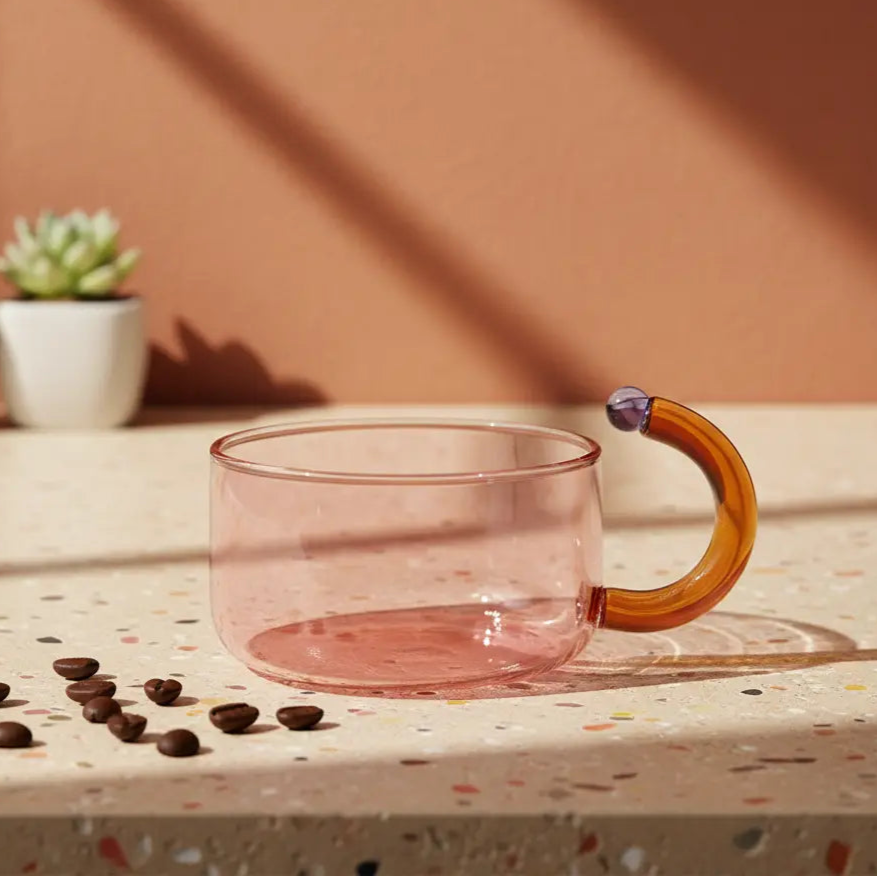 Pink glass mug with a wooden handle on a speckled surface with coffee beans and plants in the background.
