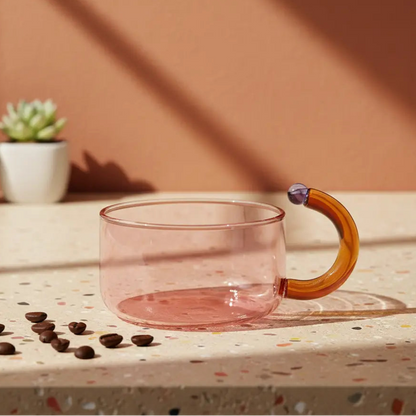 Pink glass mug with a wooden handle on a speckled surface with coffee beans and plants in the background.