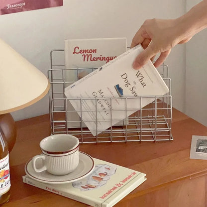 Hand placing a book into a wire bookshelf on a wooden surface with a cup and lamp.
