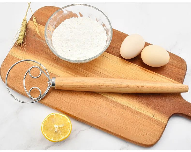 Wooden cutting board with flour, eggs, a lemon, and a whisk on a white background