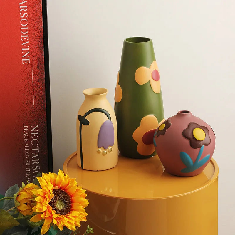 Three colorful vases on a yellow surface with a sunflower and a book in the background.