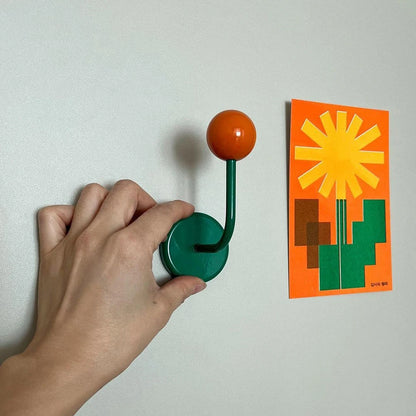 Hand using a green hook with an orange ball to hang a sun-shaped card on a white wall.