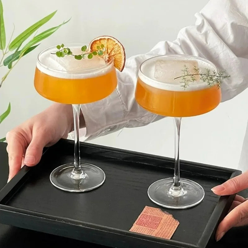 Two orange cocktails with garnishes on a black tray, held by a person wearing a white shirt.
