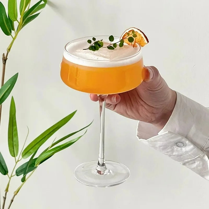 Hand holding a cocktail with an orange slice and green garnish, surrounded by green leaves on a light background