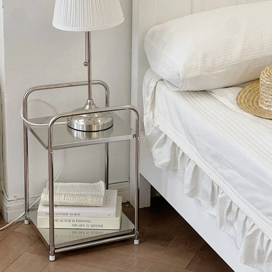 A metal trolley cart with wheels used as a bedside table. One shelf contains books and another a lamp 