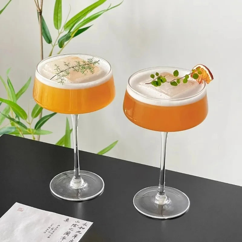 Two cocktails with orange liquid and white foam on a black surface with green plants in the background.