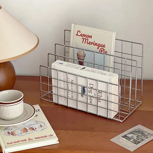 Metal wire magazine rack on a wooden surface with books and a lamp.