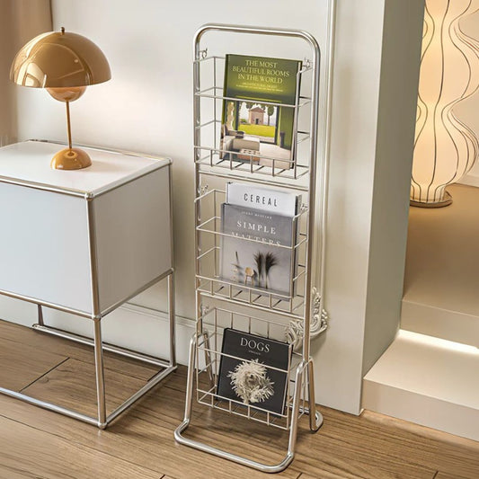 Metal magazine rack with books on a wooden floor next to a white cabinet and lamp.
