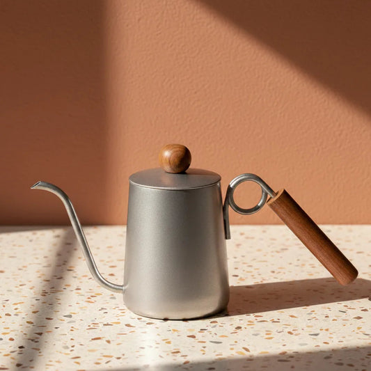 Stainless steel gooseneck kettle with wooden handle and lid on a terrazzo countertop.