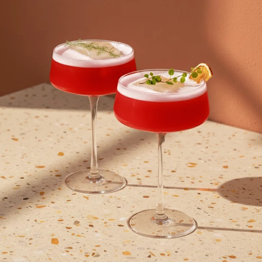 Two red cocktails with white foam and garnishes on a terrazzo countertop.