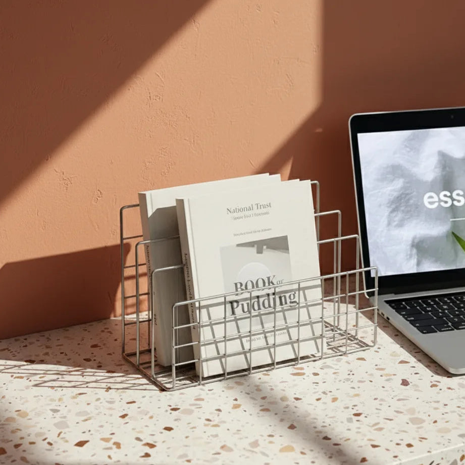 Laptop on a terrazzo desk with a wire magazine holder containing books against a peach wall.