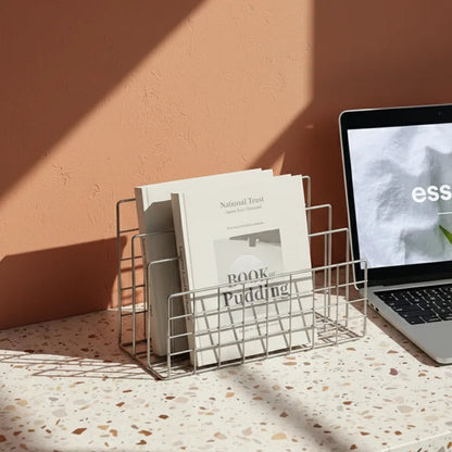 Laptop on a terrazzo desk with a wire magazine holder containing books against a peach wall.