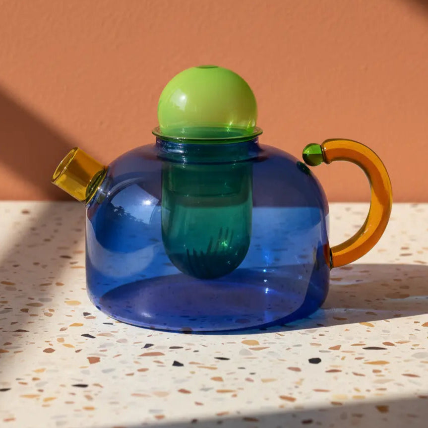 Colorful glass teapot with a green lid and amber handle on a terrazzo surface.
